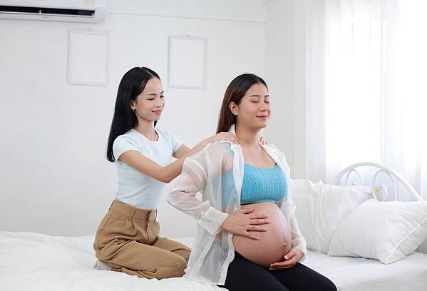 Caring female friend massaging shoulder of a pregnant woman to relieve body aches and discomfort during pregnancy on a bed.